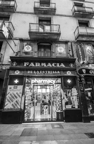 An exterior view of an old pharmacy building with vintage signage. The entrance features large glass doors displaying the word 'pharmacy' and posters on either side. The building has multiple storeys with balconies on the upper levels.