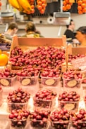A vibrant display of fresh cherries in plastic containers and cardboard boxes on a market stall. Ripe bananas and hanging clusters of tomatoes can be seen in the background, with the blurred figures of people behind the stall.