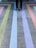A person stands on a concrete surface with colorful stripes arranged in a geometric pattern. The stripes include shades of green, blue, purple, and orange, creating a vibrant visual effect.