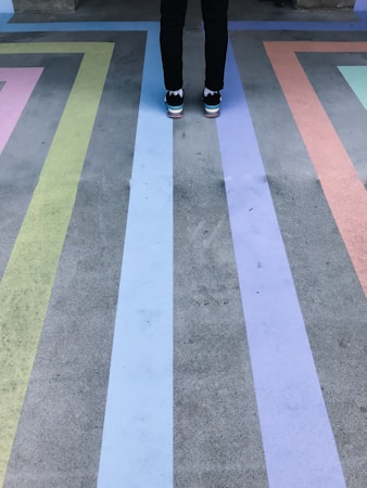 A person stands on a concrete surface with colorful stripes arranged in a geometric pattern. The stripes include shades of green, blue, purple, and orange, creating a vibrant visual effect.