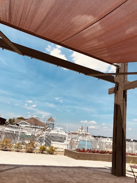 A shaded outdoor area with a pergola structure. In the background, there are boats docked near a marina visible through a white fence. The sky is clear with some scattered clouds. There is a bed of plants and flowers near the fence, and some landscaping is visible along the ground.