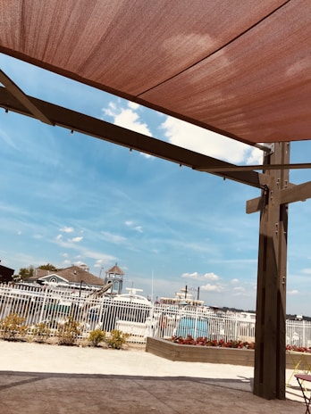 A shaded outdoor area with a pergola structure. In the background, there are boats docked near a marina visible through a white fence. The sky is clear with some scattered clouds. There is a bed of plants and flowers near the fence, and some landscaping is visible along the ground.
