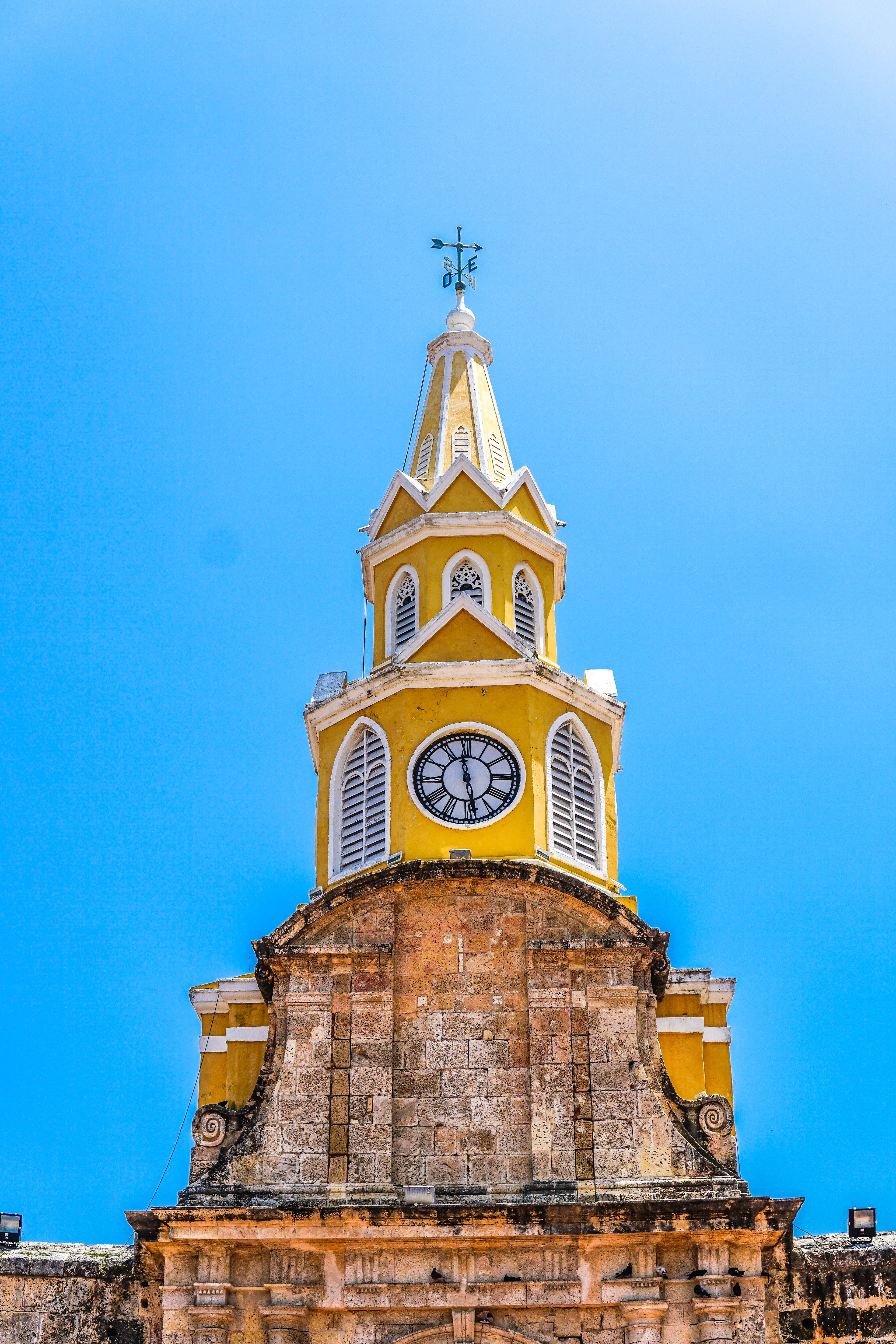 The yellow and white clock tower of Cartagena's Torre del Reloj, the main gate into the walled city, rising above its weathered coral-stone arch against a deep blue Caribbean sky.