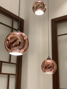 Three spherical pendant lights with a reflective copper finish, suspended from the ceiling. The reflections are detailed and include an interior staircase, wooden frames, and glass panels. The warm metallic sheen creates intriguing distortions of the surroundings.