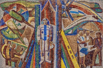 A vibrant mosaic artwork features a central figure in a space suit, surrounded by abstract representations of space exploration elements, including rockets and satellites. On the left, individuals are depicted working with technology, while on the right, a group is engaged in discussion or collaboration. The composition uses bold, geometric patterns and vivid colors to convey a futuristic theme.