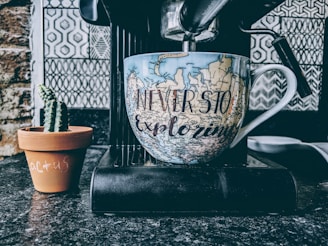 A large coffee mug with a map design and the phrase 'Never Stop Exploring' is positioned on a coffee machine, with a small potted cactus next to it. The background features a patterned tile wall.