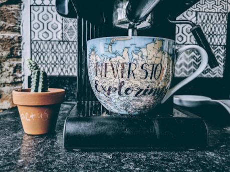 A large coffee mug with a map design and the phrase 'Never Stop Exploring' is positioned on a coffee machine, with a small potted cactus next to it. The background features a patterned tile wall.