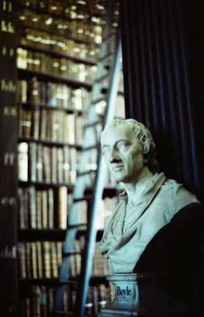 A marble bust of a historical figure is prominently displayed in front of a large bookshelf filled with numerous old, leather-bound books. A ladder leans against the shelf, suggesting a library setting.