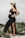 A pregnant woman stands confidently outdoors, wearing a black dress with a high slit and a straw hat. Her hands rest on her belly, and a serene expression graces her face. The setting appears to be a natural environment with greenery and sand.