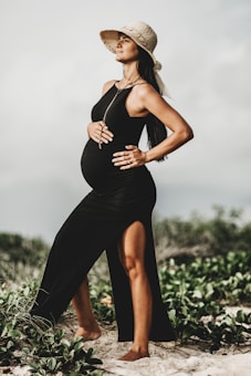 A pregnant woman stands confidently outdoors, wearing a black dress with a high slit and a straw hat. Her hands rest on her belly, and a serene expression graces her face. The setting appears to be a natural environment with greenery and sand.