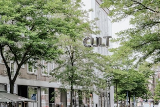A street scene with several buildings in the background, partially obscured by lush green trees. The word 'CULT' appears prominently in large letters, likely as part of a sign. The scene captures a mix of urban architecture and natural foliage.