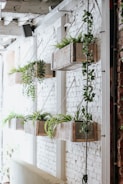 Several wooden planters containing various green plants are suspended on a white brick wall. The planters hang in a staggered line, and vines of ivy extend downward. The rustic aesthetic contrasts with the industrial feel of the surrounding structure, creating a natural yet urban ambiance.