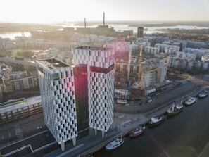 An aerial view of a modern cityscape featuring a prominent checkered building with reflective windows and an open space around it. The structure stands near a harbor with several boats docked along the water's edge. Surrounding the building are construction cranes and various urban buildings, creating a sense of ongoing development. The sun casts a warm glow over the scene, with distant smokestacks visible against the skyline.