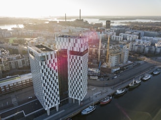 An aerial view of a modern cityscape featuring a prominent checkered building with reflective windows and an open space around it. The structure stands near a harbor with several boats docked along the water's edge. Surrounding the building are construction cranes and various urban buildings, creating a sense of ongoing development. The sun casts a warm glow over the scene, with distant smokestacks visible against the skyline.