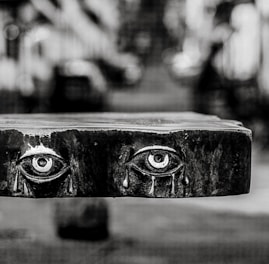 A black and white photograph of a wooden panel with realistically carved eyes, each shedding tears. The background is blurred, creating a shallow depth of field effect, placing emphasis on the intricate details of the carving.