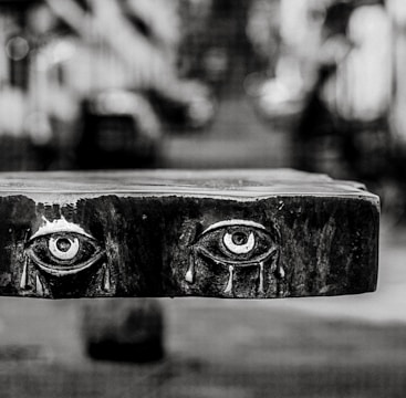 A black and white photograph of a wooden panel with realistically carved eyes, each shedding tears. The background is blurred, creating a shallow depth of field effect, placing emphasis on the intricate details of the carving.