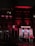 A nighttime street scene featuring a building with bright red neon lights. The building has signage with the words 'Moulin Rouge' and images related to an erotic nightclub and live theater. Two people are walking past the entrance carrying umbrellas, suggesting a rainy environment.