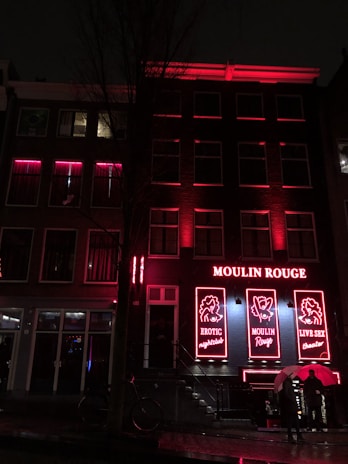 A nighttime street scene featuring a building with bright red neon lights. The building has signage with the words 'Moulin Rouge' and images related to an erotic nightclub and live theater. Two people are walking past the entrance carrying umbrellas, suggesting a rainy environment.
