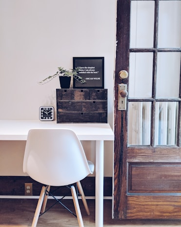 A minimalist room with a clean, white desk and a modern chair. There's a small black digital clock on the desk displaying the time '9:45'. A small black potted plant and a framed quote by Oscar Wilde are placed on a rustic wooden organizer. A wooden door with glass panes stands to the right.