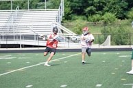 Two lacrosse players are engaged in an action-packed game on a grass field. One player in a red vest with the number 61 is holding a lacrosse stick and is in motion. The other player wears a white shirt with red markings and runs close behind. The background features empty bleachers and greenery.