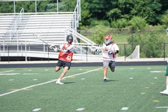 Two lacrosse players are engaged in an action-packed game on a grass field. One player in a red vest with the number 61 is holding a lacrosse stick and is in motion. The other player wears a white shirt with red markings and runs close behind. The background features empty bleachers and greenery.