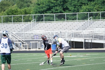 A lacrosse game in progress on a field with two players actively engaged in the play while wearing helmets and protective gear. Another player stands nearby, also wearing a uniform. The field is lined, indicative of a standard sports field, with bleachers in the background under a clear sky.