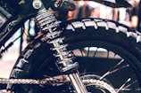 Close-up of a motorcycle rear wheel and suspension. The shiny metal coil spring and suspension system are prominently featured against the rugged tire. The image highlights the detailed engineering and mechanical components of the motorcycle.