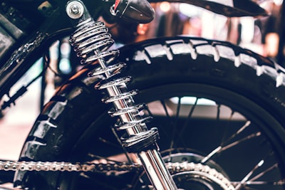 Close-up of a motorcycle rear wheel and suspension. The shiny metal coil spring and suspension system are prominently featured against the rugged tire. The image highlights the detailed engineering and mechanical components of the motorcycle.