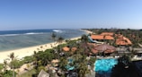 A scenic view of a beachfront resort with lush greenery, palm trees, and turquoise water. The sandy beach stretches along the coastline with gentle waves lapping against it. Several traditionally styled buildings with triangular roofs are visible amidst the tropical foliage.