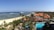 A scenic view of a beachfront resort with lush greenery, palm trees, and turquoise water. The sandy beach stretches along the coastline with gentle waves lapping against it. Several traditionally styled buildings with triangular roofs are visible amidst the tropical foliage.