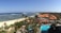 A scenic view of a beachfront resort with lush greenery, palm trees, and turquoise water. The sandy beach stretches along the coastline with gentle waves lapping against it. Several traditionally styled buildings with triangular roofs are visible amidst the tropical foliage.