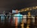 A brightly lit casino with neon signs and red lights is reflected in a river. A bridge stretches across the water, with an urban skyline visible in the background against an overcast night sky.