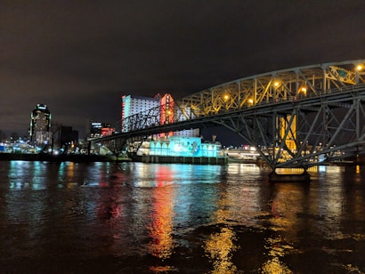A brightly lit casino with neon signs and red lights is reflected in a river. A bridge stretches across the water, with an urban skyline visible in the background against an overcast night sky.