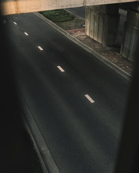 A deserted road runs alongside a large concrete bridge structure. The pavement is dark with visible cracks and faded lane markings. The bridge's support pillars rise from a brick sidewalk, with sparse greenery visible along the road's edge.