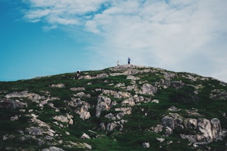 A lush green hill with rocky outcrops stretches under a wide, partly cloudy sky. Three small figures are visible at the top of the hill, suggesting a sense of achievement or exploration.