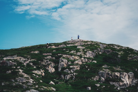 A lush green hill with rocky outcrops stretches under a wide, partly cloudy sky. Three small figures are visible at the top of the hill, suggesting a sense of achievement or exploration.