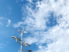 A wind turbine and two solar panels are positioned against a backdrop of a bright blue sky with scattered white clouds.