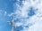 A wind turbine and two solar panels are positioned against a backdrop of a bright blue sky with scattered white clouds.