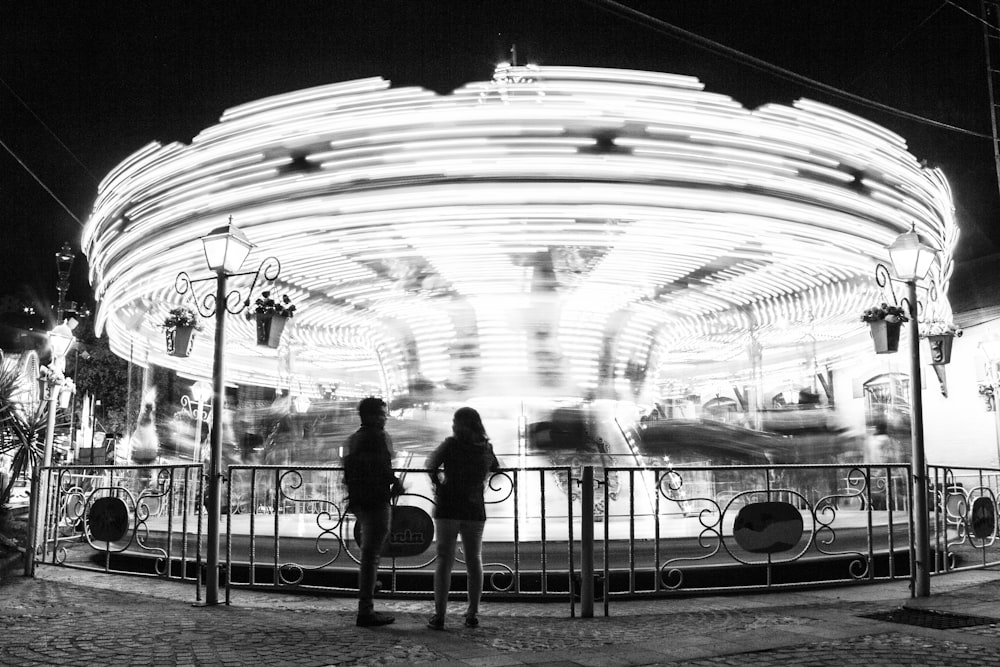 grayscale photo of man and woman standing near carousel