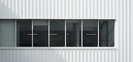 A modern facade featuring a row of tall rectangular windows set into a white corrugated metal wall. Inside the windows, linear lights are visible, hinting at an industrial or commercial interior space. The exterior surface is clean and minimalist in design.