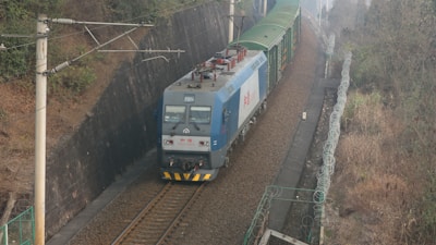 A blue freight train travels along a curving track, surrounded by a wire fence and dense greenery on both sides. The locomotive has a flat design with visible pantographs on top, indicating it is electric. The scene conveys a sense of motion and industrial activity as the train moves through a rural area.