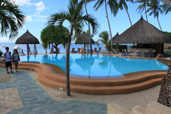 A tranquil outdoor pool area with a clear blue swimming pool surrounded by thatched-roof huts and palm trees. Two people walk along a tiled pathway beside the pool. The clear sky and distant view of the ocean create a serene and tropical atmosphere.