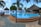 A tranquil outdoor pool area with a clear blue swimming pool surrounded by thatched-roof huts and palm trees. Two people walk along a tiled pathway beside the pool. The clear sky and distant view of the ocean create a serene and tropical atmosphere.