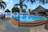A tranquil outdoor pool area with a clear blue swimming pool surrounded by thatched-roof huts and palm trees. Two people walk along a tiled pathway beside the pool. The clear sky and distant view of the ocean create a serene and tropical atmosphere.