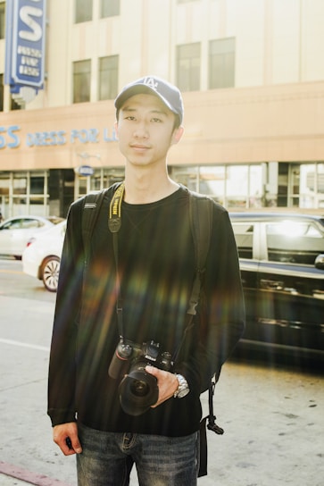 Portrait of Neil Pollick holding a camera, standing in a sunlit urban street.