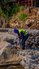A person wearing a blue hood and yellow safety vest is working near a rocky embankment. Scrub and greenery grow on the hillside above the rocks, while metal wire meshes hold the stones in place. A red and black rail fence runs along the top, and various construction equipment is visible in the background.