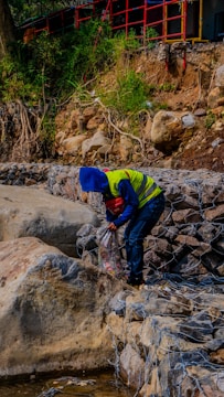 A person wearing a blue hood and yellow safety vest is working near a rocky embankment. Scrub and greenery grow on the hillside above the rocks, while metal wire meshes hold the stones in place. A red and black rail fence runs along the top, and various construction equipment is visible in the background.