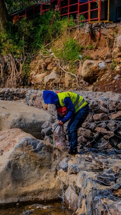 A person wearing a blue hood and yellow safety vest is working near a rocky embankment. Scrub and greenery grow on the hillside above the rocks, while metal wire meshes hold the stones in place. A red and black rail fence runs along the top, and various construction equipment is visible in the background.