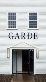 A white building with vertical siding and two rectangular windows above an entrance. The building has the word 'GARDE' in large black letters on the facade. The entrance features a glass door and decorative lamps on either side. The number '2280' is visible near the door.