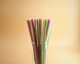 A collection of colorful straws stands upright in a clear glass against a warm, beige background. The straws vary in hues, including purple, green, and beige, and are arranged closely together.